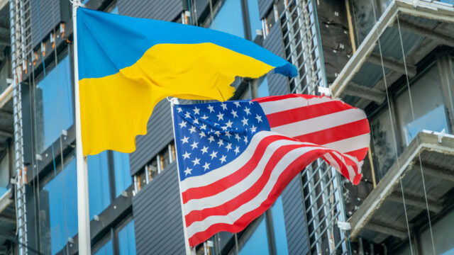 A Ukrainian flag and an American flag fly against a blue sky in front of a building