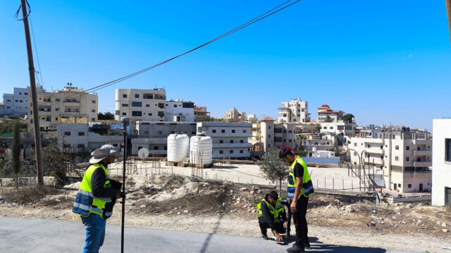 Three men in yellow vests standing on the street in a city holding survey equipment