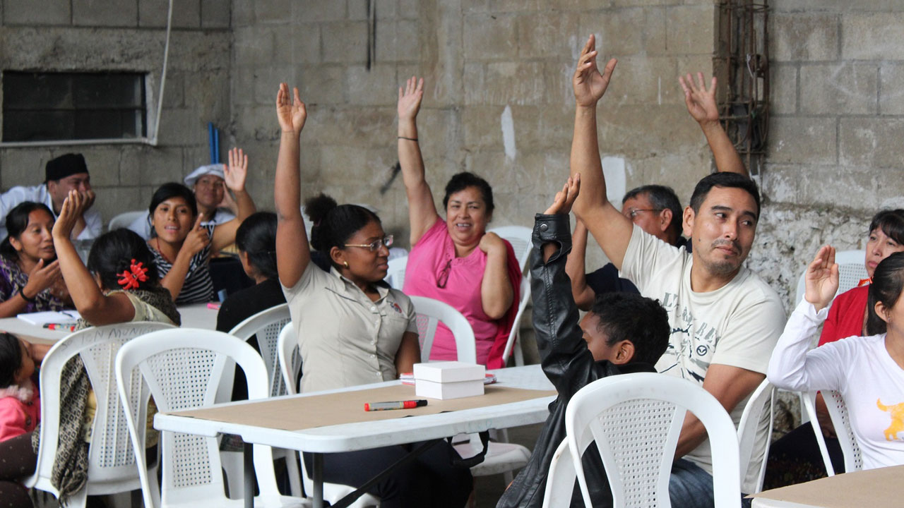 A group of people sitting around tables indicates their vote with their raised hands